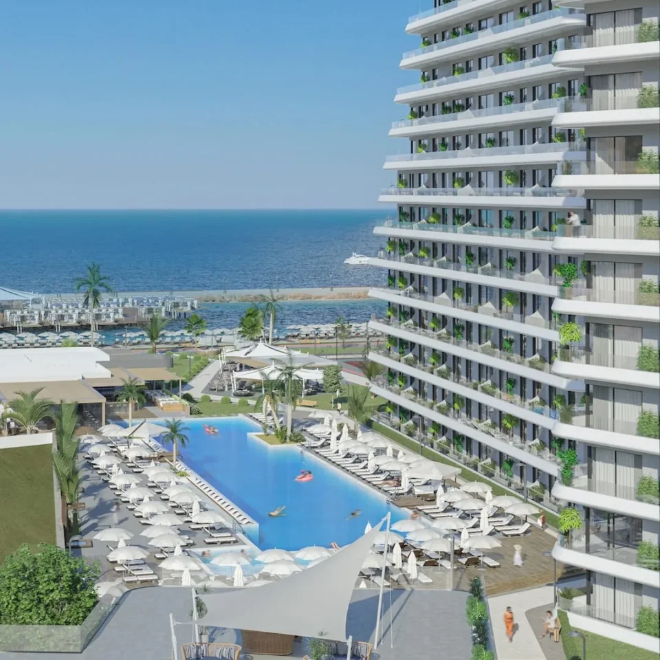 Alpcans A high-rise beachfront resort features a large pool surrounded by lounge chairs and umbrellas. The building has balconies with greenery, and the beach is visible in the background.