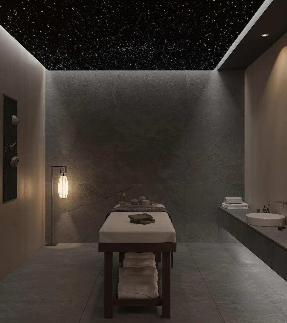 Alpcans A modern, minimalist spa room with a massage table, soft lighting, and a dark ceiling with small light fixtures resembling stars. Towels and spa supplies are neatly arranged on and around the table.