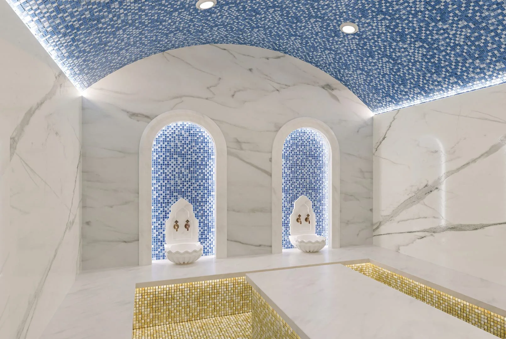 Alpcans A modern Turkish bath with marble walls, a blue mosaic tile ceiling, and two alcoves with fountains.